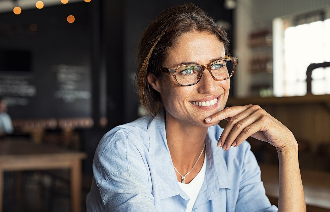 A woman wearing glasses and a blue shirt smiles, sitting in a dimly lit cafe.