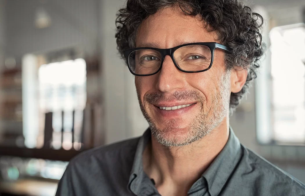 A man with curly hair and glasses smiles at the camera, wearing a gray shirt. The background is softly blurred.