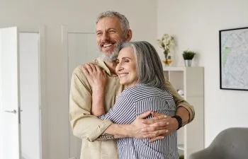 An older couple embraces and smiles in a modern living room with a map on the wall.