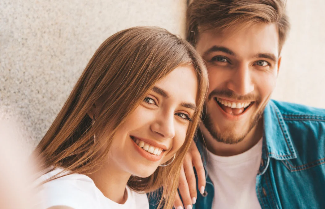 A smiling man and woman take a close-up selfie against a light-colored wall.